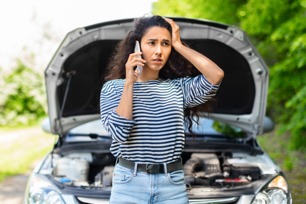 Nervous young woman standing by open car hood and calling to auto service, got broken at highway on a way to countryside. Female drive having phone conversation with friends, asking for help with auto